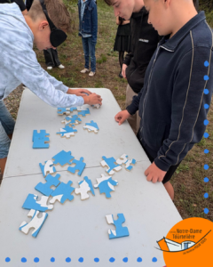 Un groupe d'élèves du Lycée Notre-Dame de la Tourtière participe à une activité de sensibilisation. Un élève est aveuglé par un bandeau et tente de jouer aux échecs sur une table, guidé par ses camarades.